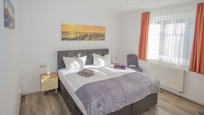 Ein modernes Schlafzimmer mit Bett, Nachttisch, Sessel, Gemälde und einem Fenster mit Vorhängen.