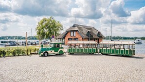 Eine grüne und weiße Straßenbahn an einem Seeufer, ein Gebäude mit Strohdach und Boote im Hintergrund.