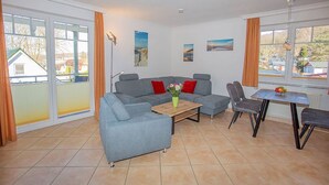 Ein Wohnzimmer mit grauem Sofa, einem Holztisch und einem Esstisch mit Stühlen. Große Fenster mit Vorhängen und Kunstwerke an den Wänden.