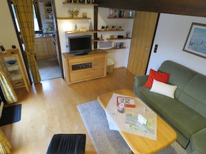Un soggiorno con un divano verde, un tavolino in legno con dei documenti, una TV su un mobile in legno e una cucina sullo sfondo.