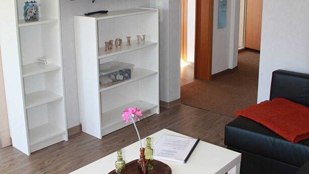 Una sala de estar moderna con un televisor de pantalla plana, estanterías blancas, una mesa de centro con un jarrón y flores, y un sofá negro con una manta roja.