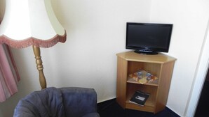 Een kamer met een grijze fauteuil, een houten tv-meubel met een flatscreen televisie, en een lamp met een witte kap en roze accenten.