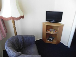 Une pièce avec un fauteuil gris, un meuble TV en bois sur lequel est posée une télévision à écran plat, et une lampe avec un abat-jour blanc et des bordures roses.