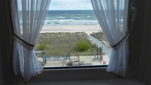 Uitzicht vanuit een raam met dunne gordijnen, op een strand met een houten looppad en een hangmat.