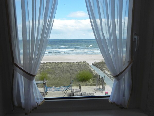 A view from a window with sheer curtains, overlooking a beach with a wooden walkway and a hammock.