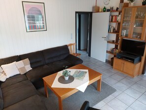 A living room with a dark brown sofa, a wooden coffee table with a map, a television on a wooden stand, and a bookshelf with various items.