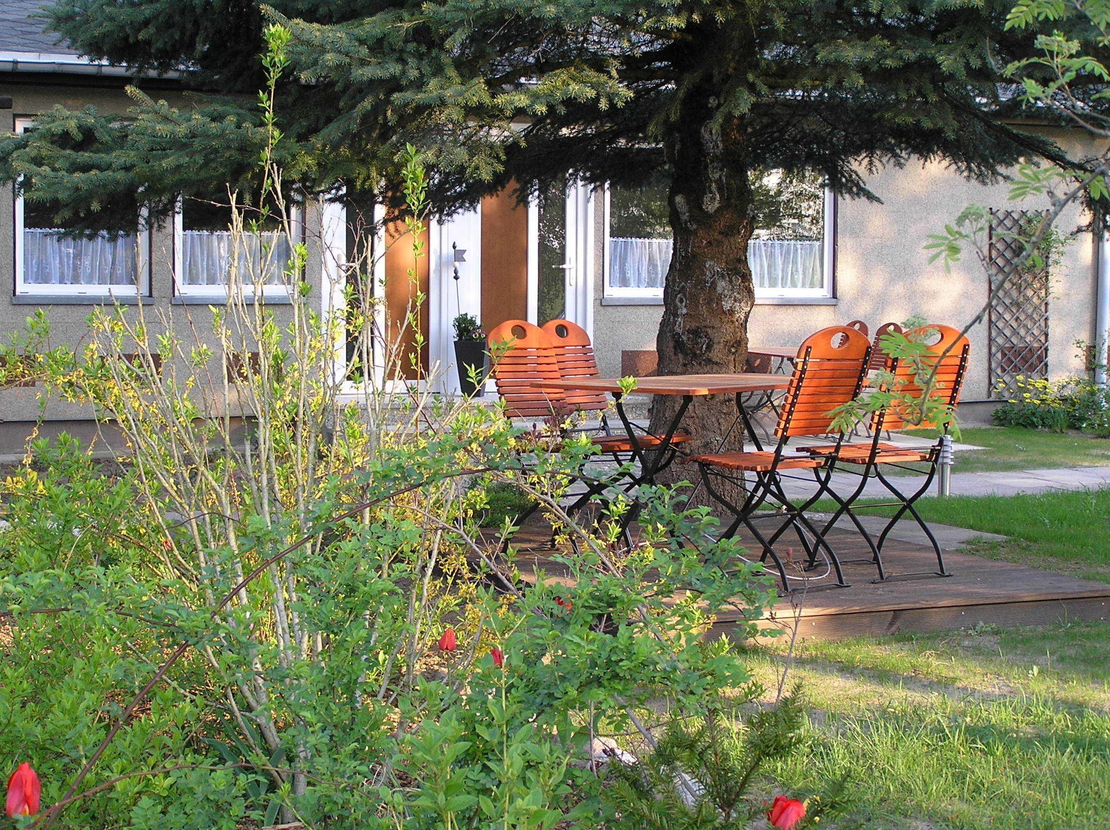A patio with a table and four chairs, surrounded by greenery and a tree.