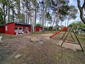 Une aire de jeux avec un toboggan rouge et des balançoires, entourée d’arbres et de bâtiments rouges.