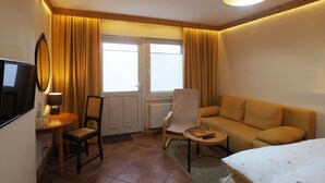 Habitación de hotel compacta con sofá, una silla, una mesita, televisor y ventana con cortinas.