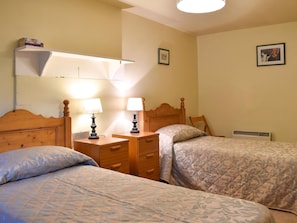 A bedroom with two beds, bedside tables with lamps, a shelf with books, and a radiator.
