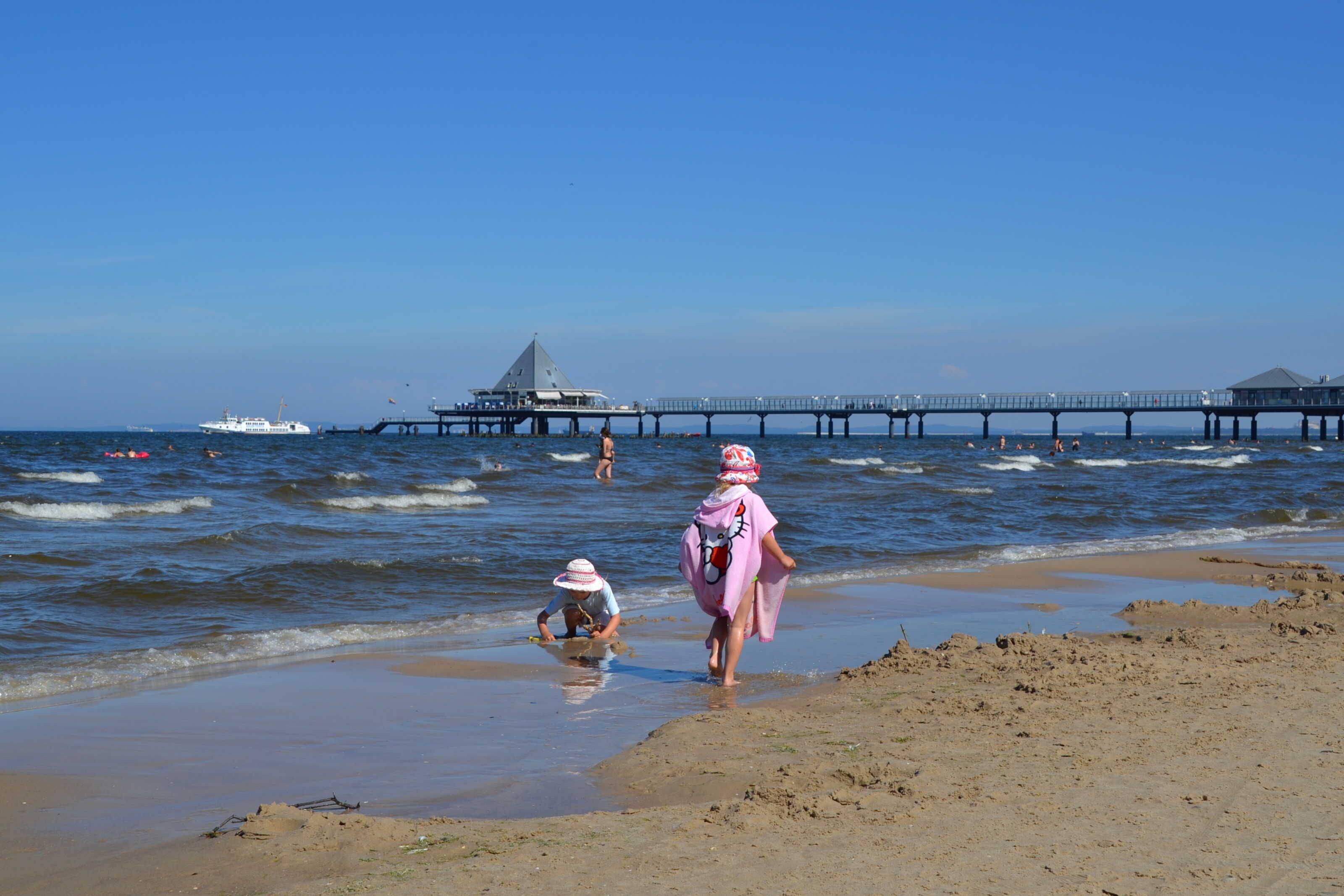 Una playa con un muelle que se adentra en el agua, un barco y gente cerca de la orilla.