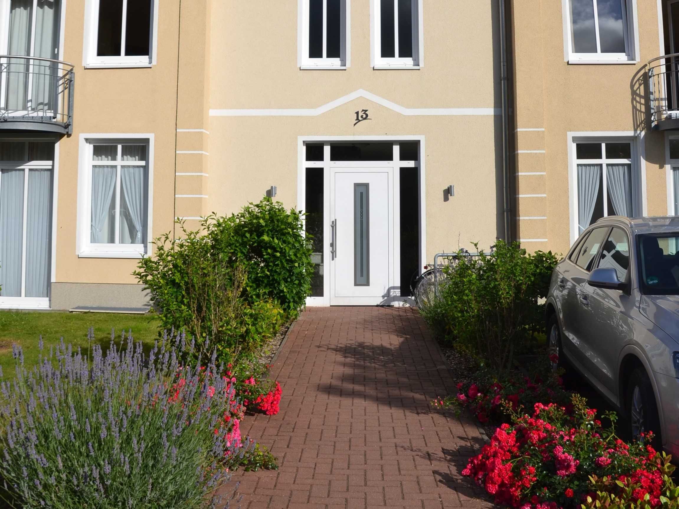 A building with a beige facade, a white door, and the number 13. There are plants and a car in front.