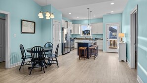 A modern kitchen with light blue walls, white cabinets, and a central island. There is a dining area with a table and chairs, and a hanging light fixture above. - Night N Gale-Close to Beach Access in Atlantic Beach, NC (Atlantic Beach)