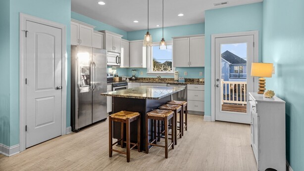 A modern kitchen with a central island, white cabinets, stainless steel appliances, and a view of a blue house through a door. - Night N Gale-Close to Beach Access in Atlantic Beach, NC (Atlantic Beach)