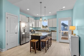 Een moderne keuken met een centraal kookeiland, witte kasten, roestvrijstalen apparatuur en uitzicht op een blauw huis door een deur.
