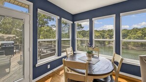 Comedor con mesa y sillas, ventanas amplias con vista a un lago y árboles, y una parrilla afuera.