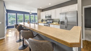 Una cocina moderna con una isla central, armarios blancos, electrodomésticos de acero inoxidable y amplios ventanales con vistas a zonas verdes.