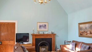 A living room with a fireplace, leather sofas, a television, and a chandelier. - Tressady Coach House (Rogart, near Dornoch)