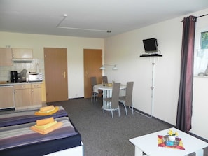 Ett kompakt hotellrum med ett litet pentry, en matplats, en tv som är monterad på väggen och ett litet bord med en vas med blommor.