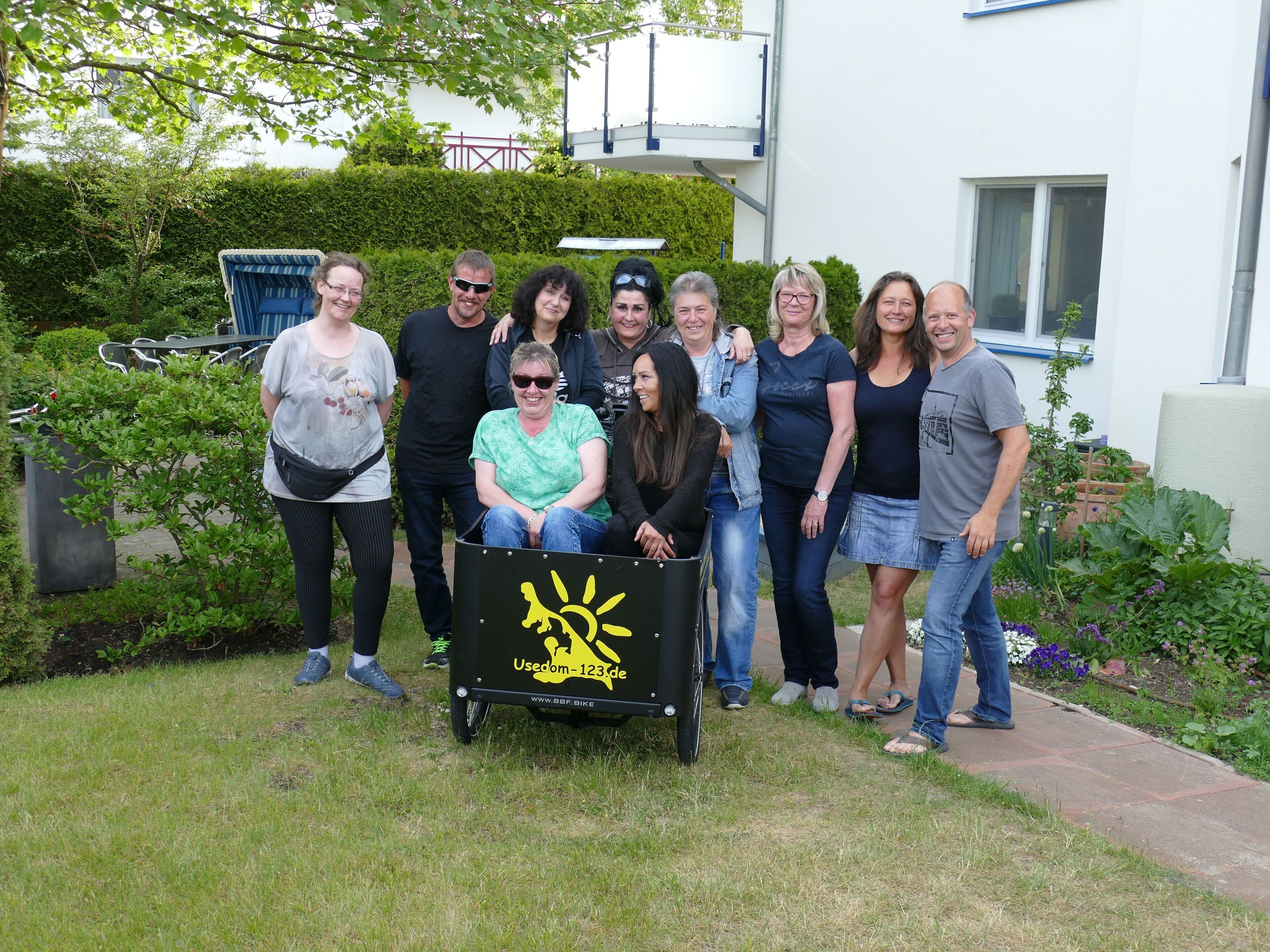 A group of people gathered around a black cart with a yellow sun design, standing in a garden with a white building in the background.