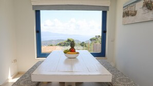 A dining table with a bowl of fruit and a view of a coastal landscape through a large window.