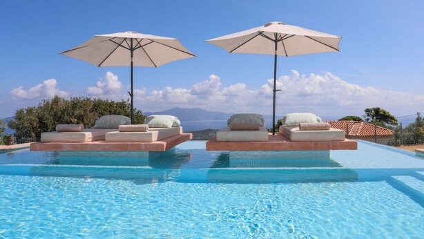 A pool with tiered seating and umbrellas, surrounded by greenery and a clear sky.