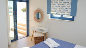 A modern hotel room with a bed, a small desk, a wicker chair, a mirror, and a window with patterned curtains.