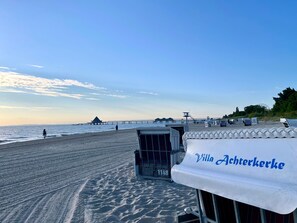 Vista da praia com uma torre salva-vidas, cadeiras de praia e uma tenda com a inscrição 'Villa Achterkerke'.