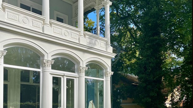 A white building with ornate columns and arched windows, a covered porch, and a well-maintained garden with white flowers and shrubs. - Apartment III: Ground floor South & Sea - Villa Achterkerke (Heringsdorf)