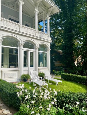 A white building with ornate columns and arched windows, a covered porch, and a well-maintained garden with white flowers and shrubs.