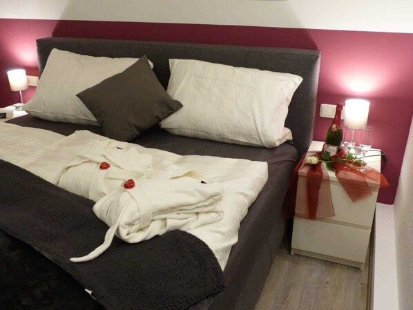 Una cama bien hecha con almohadas, una mesita de noche con una botella y vasos, y una pared con una franja roja.
