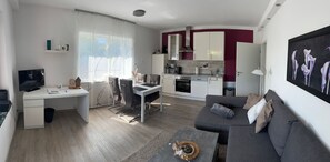 A modern living room with a grey sofa, a dining area with a white table and chairs, a kitchen with white cabinets and a red accent wall, and a TV on a white desk.