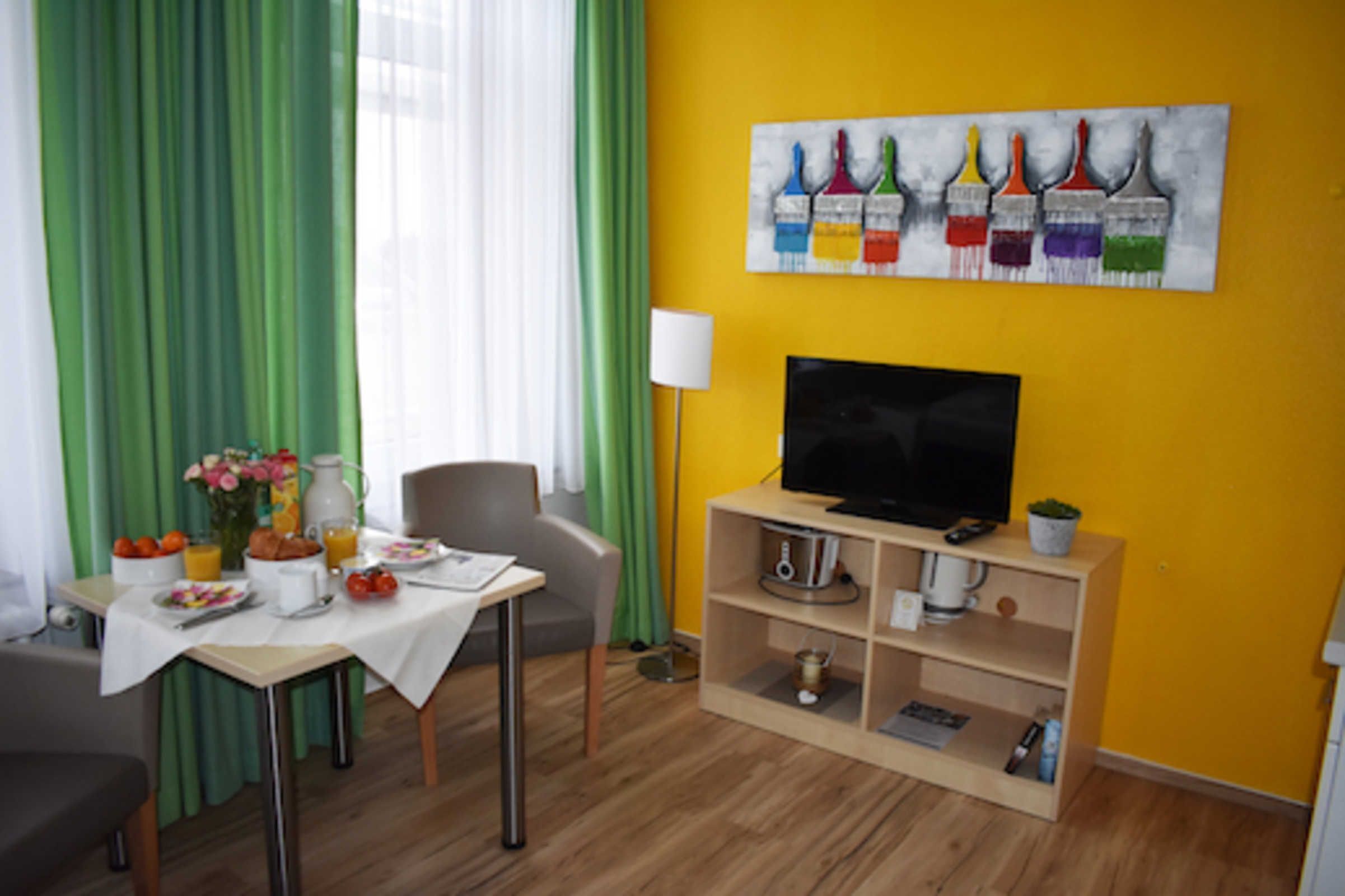 A room with a dining table set for breakfast, a television on a stand, and a painting on the wall.