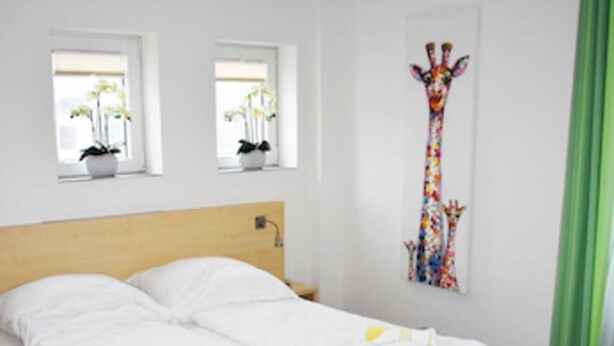Un lit bien fait, avec du linge de lit blanc, une tête de lit en bois et un protège-matelas bleu. Il y a deux fenêtres avec des plantes en pot sur les rebords, et une œuvre d’art sur le mur dont le thème est la girafe.