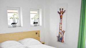 Une chambre à coucher avec un lit, deux fenêtres ornées de plantes, un tableau représentant une girafe et un rideau vert.