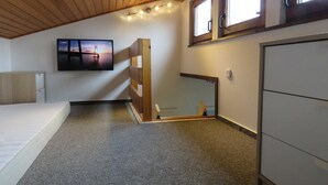 Una habitación con techo inclinado, una cama, un televisor montado en la pared, una ventana y un armario.