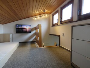 Quarto com teto inclinado, uma cama, televisão na parede, janela e guarda-roupa.