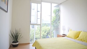 Ein Schlafzimmer mit großem Fenster, einem Bett mit gelber Bettwäsche, einem Nachttisch mit Lampe und einer Zimmerpflanze.