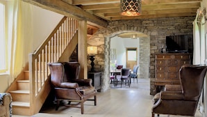 A spacious room with a wooden staircase, leather armchairs, a dining table with chairs, a stone fireplace, and a television.