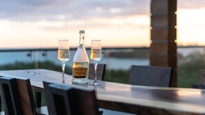 Mesa com uma garrafa e duas taças de Prosecco em um terraço com vista para a água ao pôr do sol.