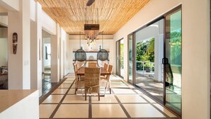 Uma área de refeições moderna com teto de madeira, uma mesa grande com cadeiras e vista para o exterior através de portas de vidro.