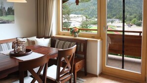 A dining area with a wooden table and chairs, a bench, a window with a view of greenery, and a hanging light fixture. - Ferienwohnung für 2 Personen mit großem Balkon, 79 m² (Inzell)