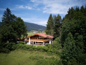 Une grande maison en bois à deux étages, avec un toit recouvert de tuiles rouges, entourée d’une forêt dense et offrant une vue sur un petit village au loin.