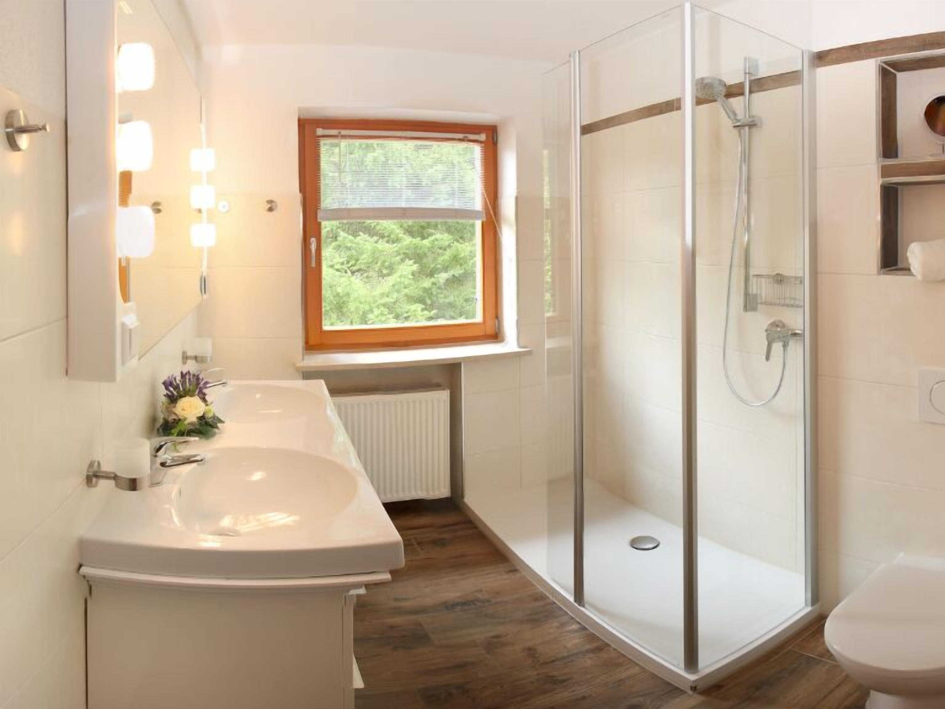 A modern bathroom with a glass-enclosed shower, a white sink, and a window with a view of greenery.