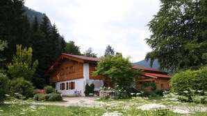 A wooden house with a sloped roof, surrounded by a garden with white flowers and trees. - Ferienwohnung für 2 Personen mit großem Balkon, 79 m² (Inzell)