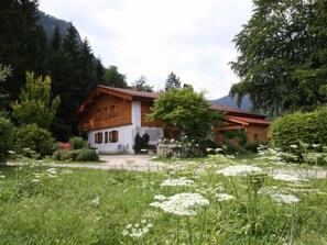 Une maison en bois avec un toit en pente, entourée d’une végétation luxuriante et d’un jardin où poussent des fleurs blanches.