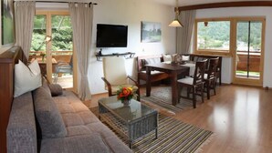 A living room with a sofa, dining table, chairs, a TV, and a view of greenery through large windows. - Ferienwohnung für 2 Personen mit großem Balkon, 79 m² (Inzell)