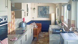 A compact kitchen with a built-in oven, a toaster, and a microwave. There is a bench with a cushion, a hanging plant, and a window with a view. - 4 bedroom accommodation in Keswick (Keswick)