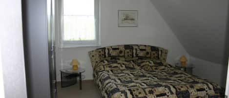 A bedroom with a bed, a window, a small table with a lamp, and a picture on the wall.