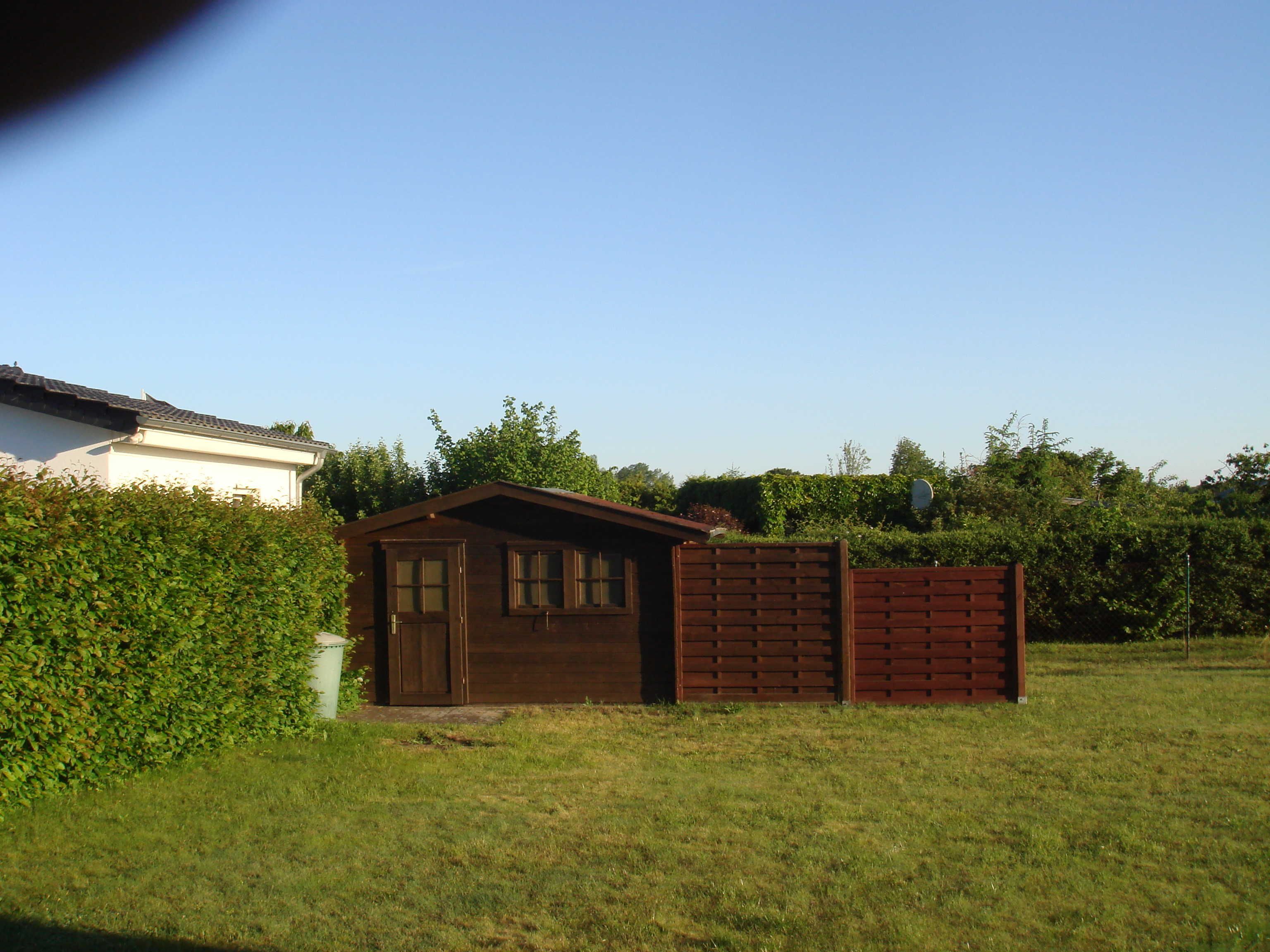 Un cobertizo de madera pequeño con puerta y ventanas, un muro de ladrillo y un seto en un patio cubierto de césped.